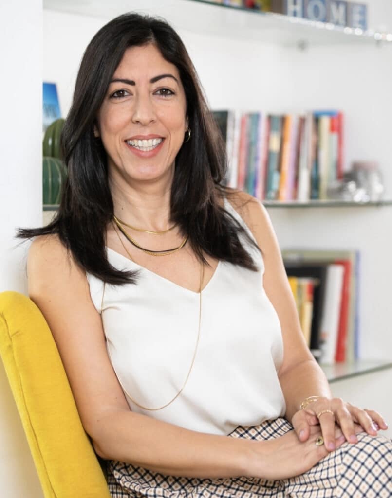 אישה עם שיער ארוך וכהה יושבת על כיסא צהוב, מחייכת, מול מדפים עם ספרים וחפצי נוי.
